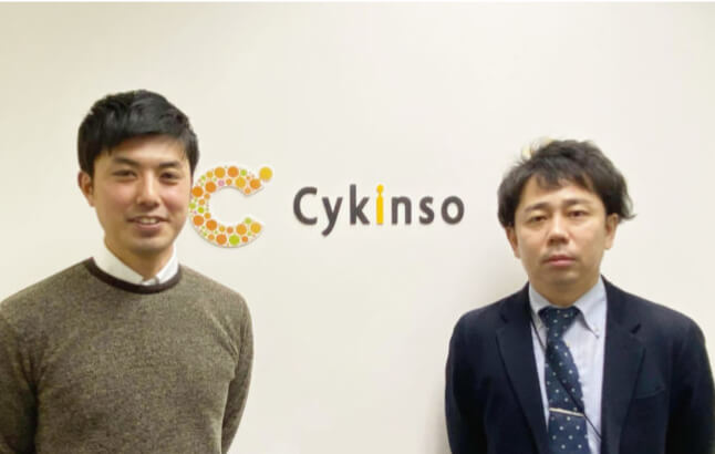 会社のロゴ前で二人の男性が並んでいる様子